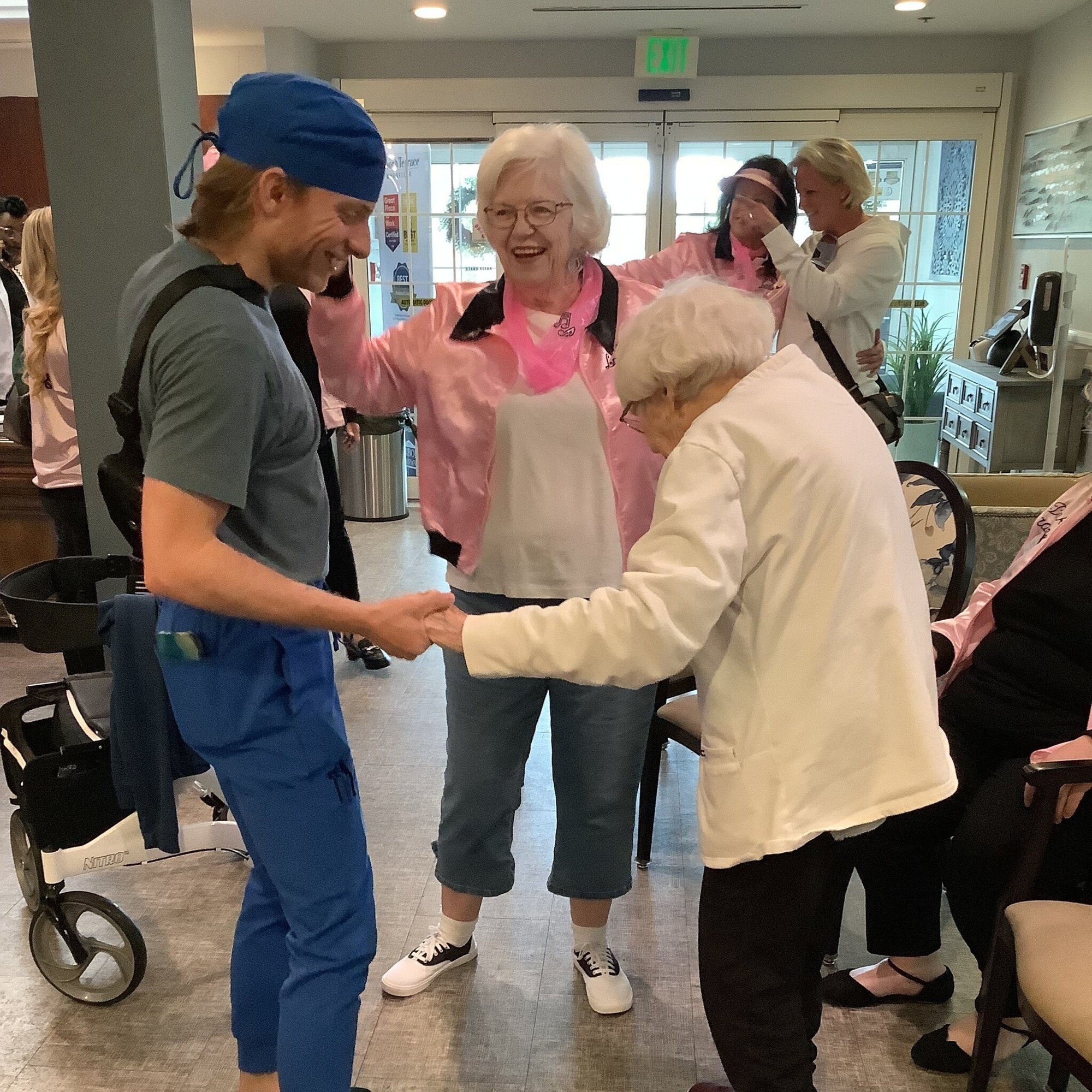 A man in scrubs dances hand in hand with a woman while another in a pink satin jacket cheers them on at Arbor Terrace Knoxville, part of social and activity-based senior living in Knoxville, TN.