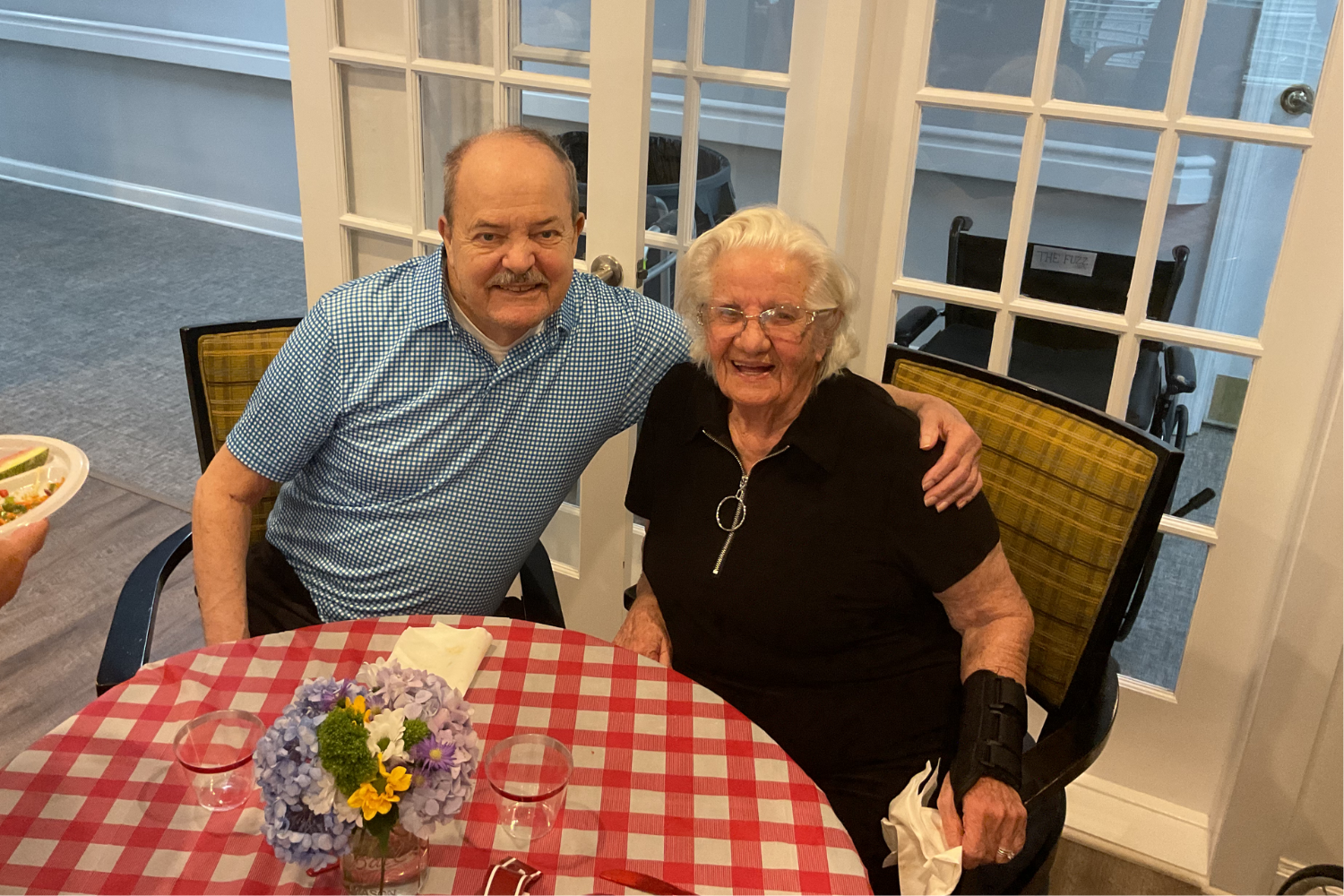A man and a woman smile side by side at a table set with a red gingham cloth and fresh flowers at Arbor Terrace Knoxville, where social and activity-based senior living in Knoxville, TN makes every shared meal feel special.