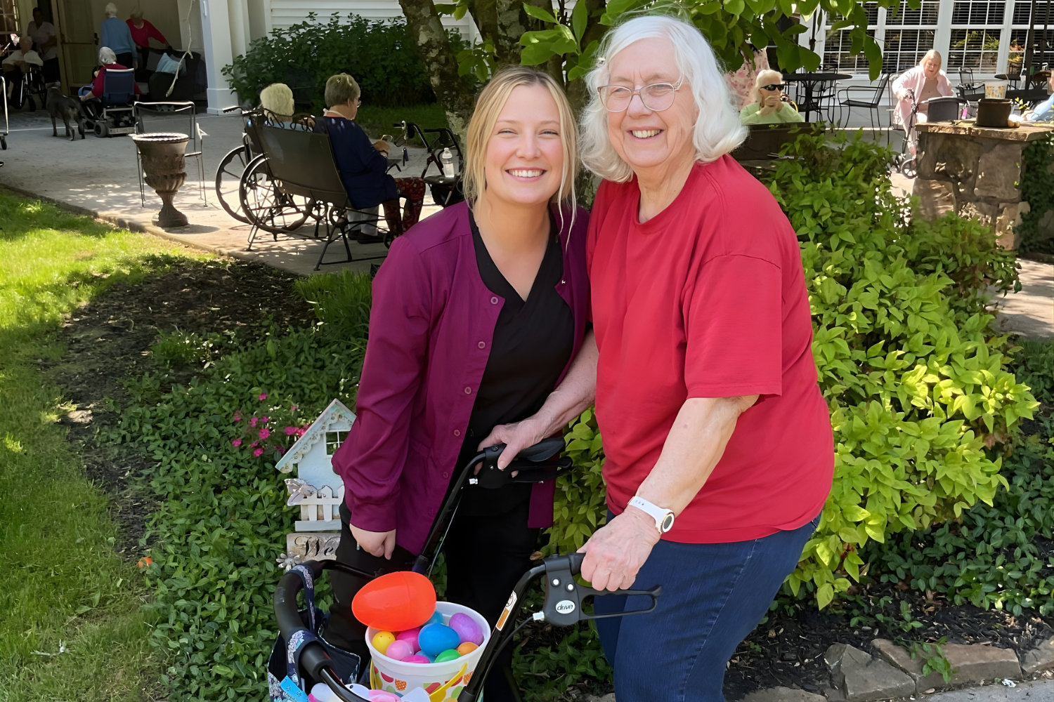A team member in scrubs and a woman with a walker smile together beside a bucket of colorful plastic eggs in the courtyard at Arbor Terrace Knoxville, part of the active senior lifestyle and senior living Knoxville, TN offers.