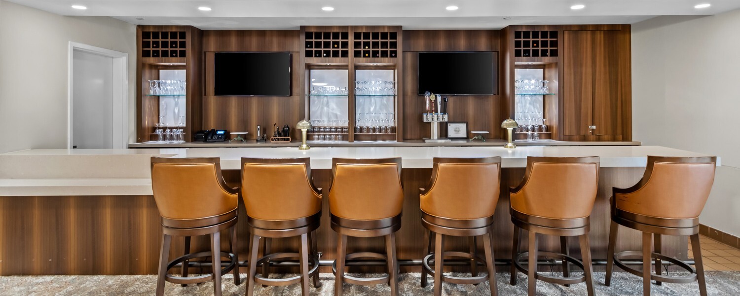 A row of cushioned bar stools lines a modern communal counter in a lounge area, with wood-paneled walls, shelving, and soft lighting, and a ‘Featured’ label displayed at the top.