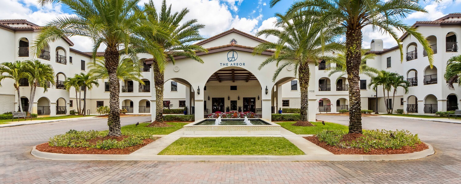 The Arbor building exterior featuring a grand arched entrance portico, white Mediterranean-style architecture with balconies, lush tropical palm trees, and a decorative fountain surrounded by manicured landscaping on a sunny day.