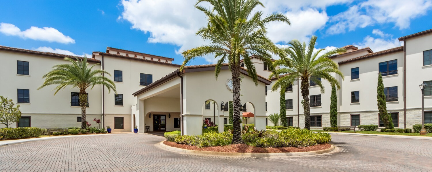 The welcoming exterior of The Arbor at Lake Worth, a senior living community in Lake Worth, FL, featuring a grand entrance surrounded by tall palm trees and lush tropical landscaping.