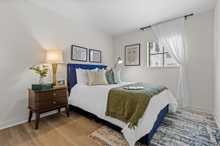 A bright, elegantly styled bedroom at this assisted living community serving Lake Worth FL, featuring a navy upholstered bed frame, crisp white bedding, a green throw blanket, warm wood flooring, framed artwork, and sheer curtains letting in soft natural light.