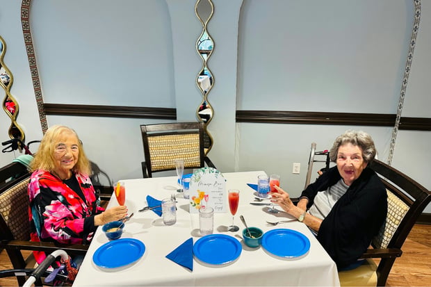 Two smiling women share a cheerful meal at a table set with crisp white linens, bright blue plates, and colorful drinks, one raising her glass in a friendly toast, a warm dining moment at one of the retirement communities Lake Worth FL offers for senior living Lake Worth Florida.