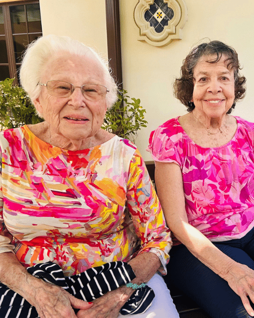 Two smiling women in bright pink floral tops sit side by side in a sunny spot framed by greenery, their warm expressions capturing the friendships that bloom in coastal senior living Palm Beach County and senior living near Lake Worth Beach.