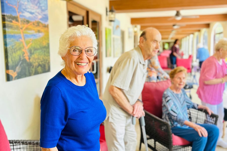 Smiling elderly woman with short white hair and glasses wearing a blue top, standing on a sunny porch at a senior living facility, with other older adults seated and socializing in the background.