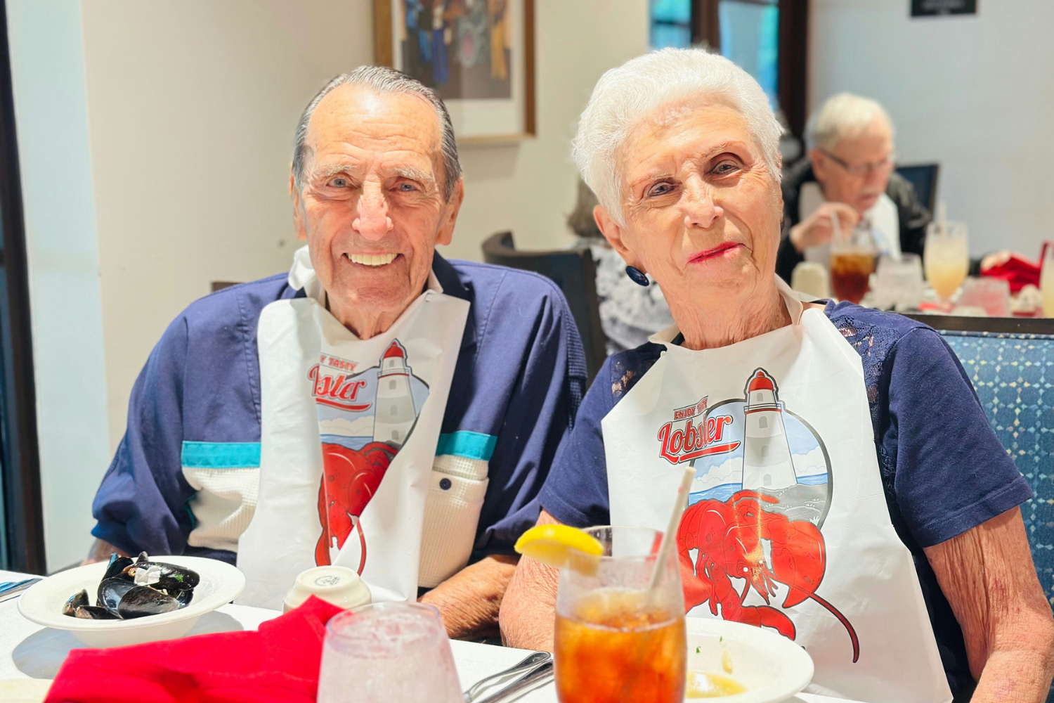 Two residents smile warmly while enjoying a festive lobster dinner, wearing themed bibs in the bright and welcoming dining room. Senior living Lake Worth FL.