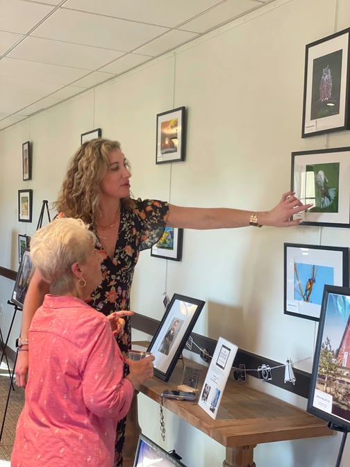 A woman in a floral dress points out framed nature photographs on a wall as a woman with white hair in a pink top looks on, sharing an art display that celebrates the active senior lifestyle Lake Worth offers at this senior community near Downtown Lake Worth.