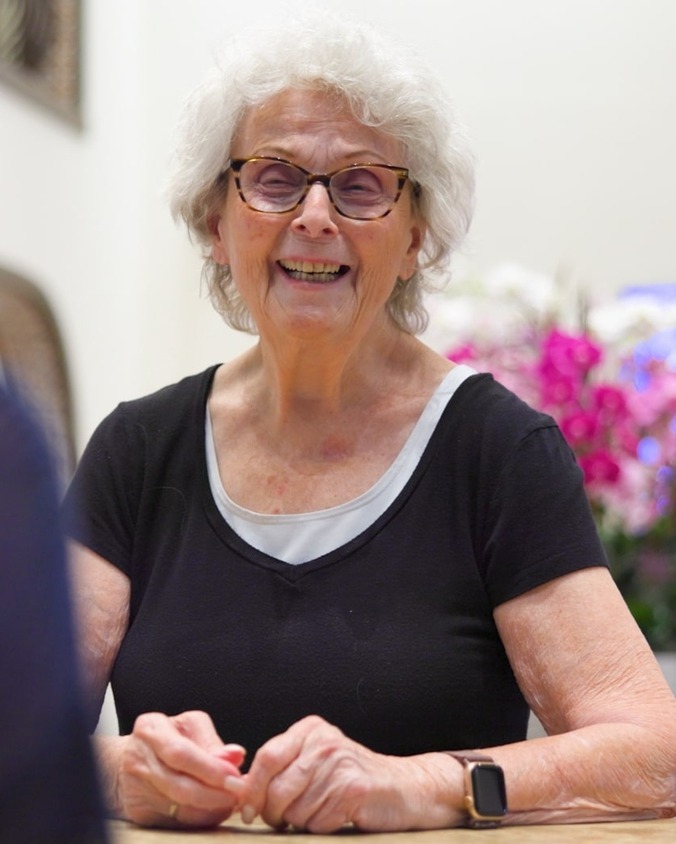  A resident with white curly hair and glasses smiling warmly while seated at a table, with colorful flowers in the background at this welcoming senior care community in Lake Worth, FL.