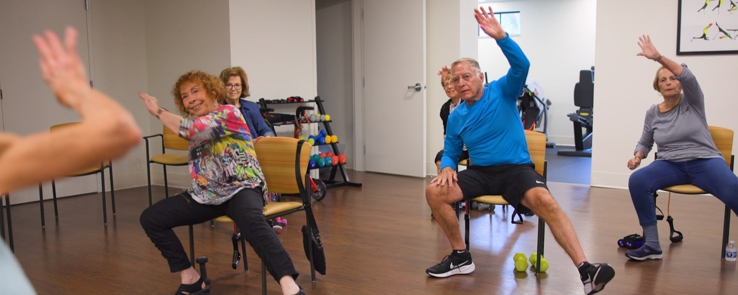 Residents enjoying a chair exercise class at this assisted living community in Lake Worth, FL, raising their arms and smiling in a bright, well-equipped fitness room with dumbbells and workout equipment nearby.