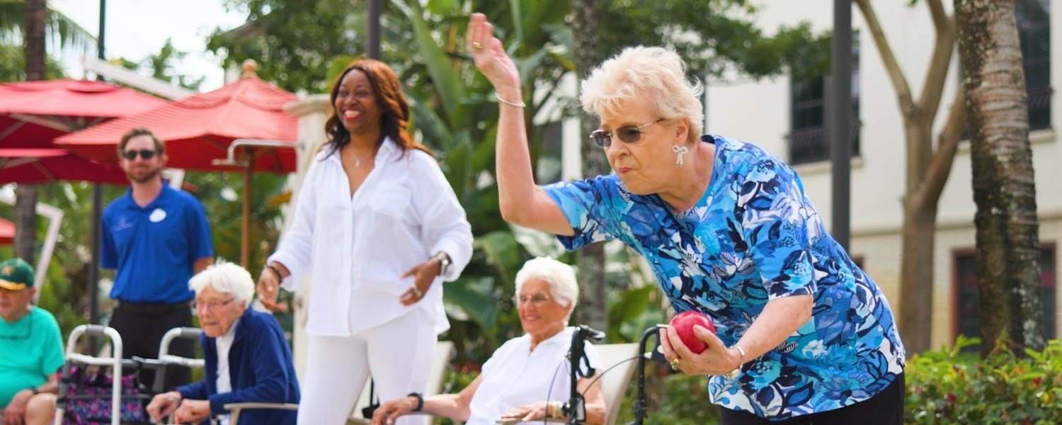A resident mid-throw during a lively bocce ball game at this senior living Lake Worth Florida community, with fellow residents and staff cheering on in a beautifully landscaped outdoor setting. 