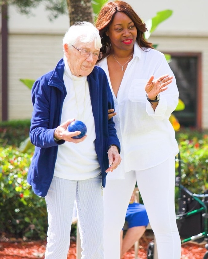 A senior lady being taught how to play bocce ball by a staff member on a sunny day at The Arbor at Lake Worth