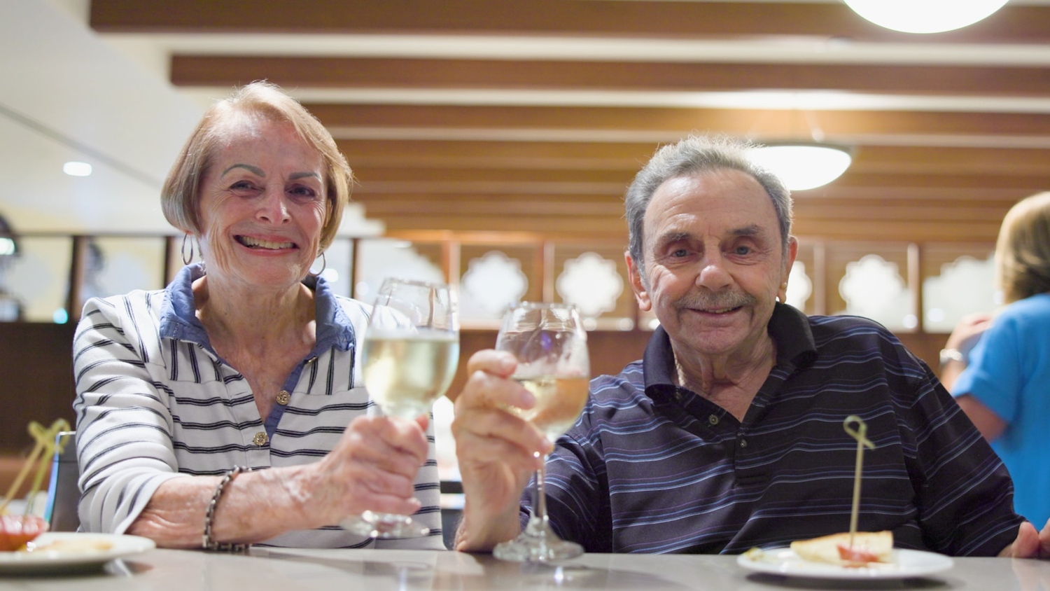 An elderly couple seated at a restaurant table smile at the camera while raising glasses of white wine in a toast, with small appetizers and warm indoor lighting in the background.