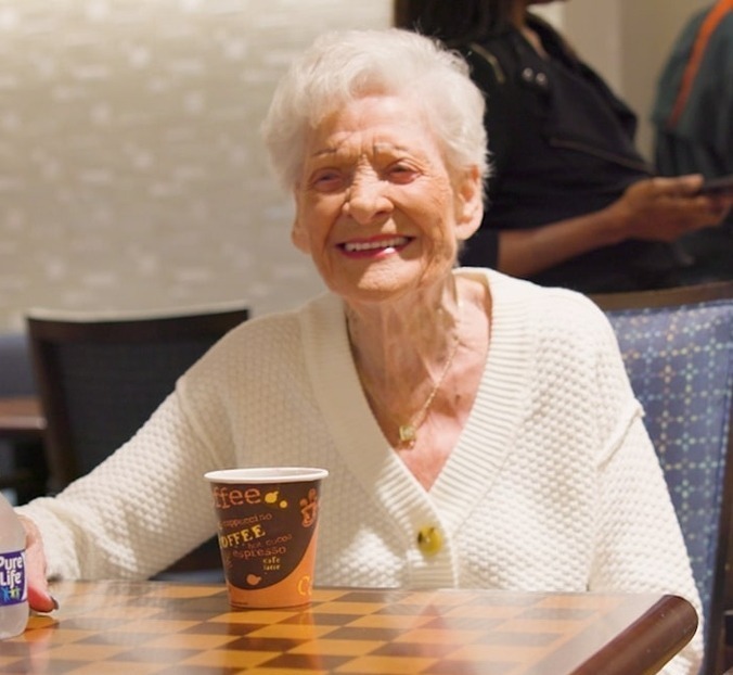 A smiling woman with white hair and a cream cardigan sits at a checkerboard table with a cup of coffee, her bright, joyful expression reflecting the welcoming spirit of retirement communities Lake Worth FL and senior living near Delray Beach.
