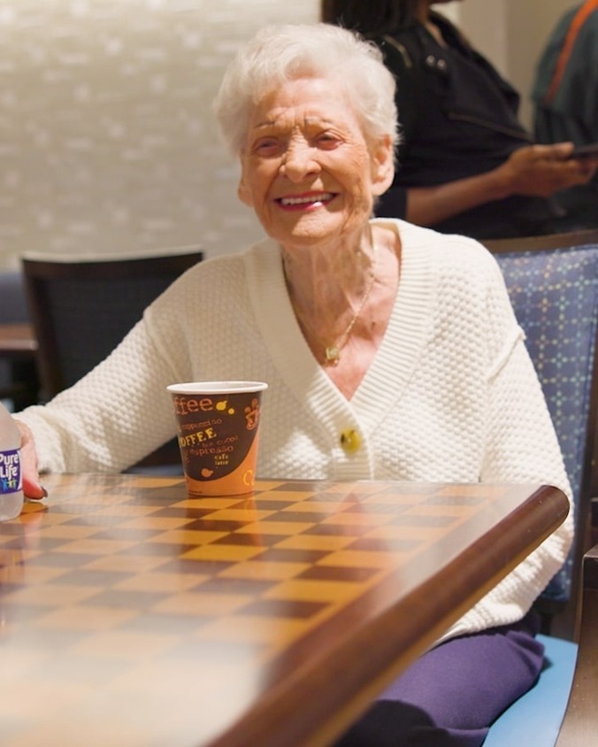 A resident smiling cheerfully while seated at a checkerboard table with a coffee cup nearby at this senior care community in Lake Worth, FL, where engaging daily activities help foster joy and connection.