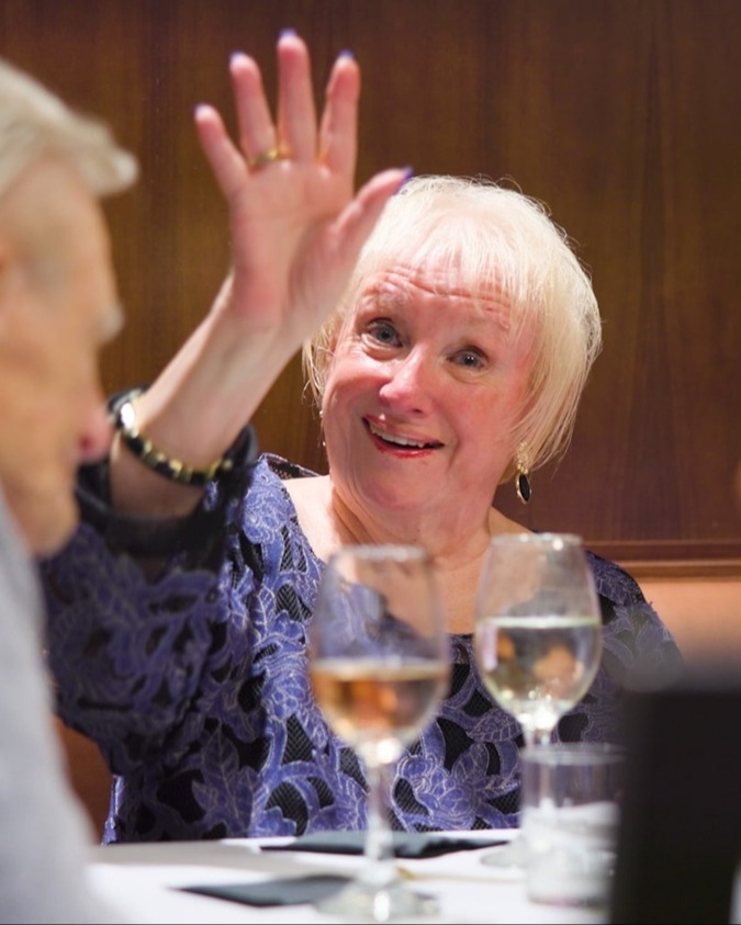 A resident laughing and gesturing expressively during a social dining experience at this senior living community in Lake Worth, Florida, with wine glasses on the table adding to the warm, welcoming atmosphere.