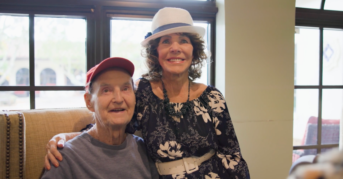A resident and a visitor sharing a joyful moment together in a bright, welcoming common area at this senior living community in Lake Worth, FL, both wearing fun hats and smiling warmly.