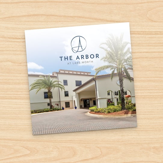 Square photo of The Arbor at Lake Worth building exterior with palm trees, paver driveway, and landscaped entrance, displayed on a printed card against a wood-grain background