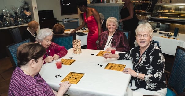 Four women gather around a white-draped table for a lively game of bingo with gold cards, black chips, and bright red drinks, a festive gathering that captures the social spirit of coastal senior living Palm Beach County at this senior community near Downtown Lake Worth.