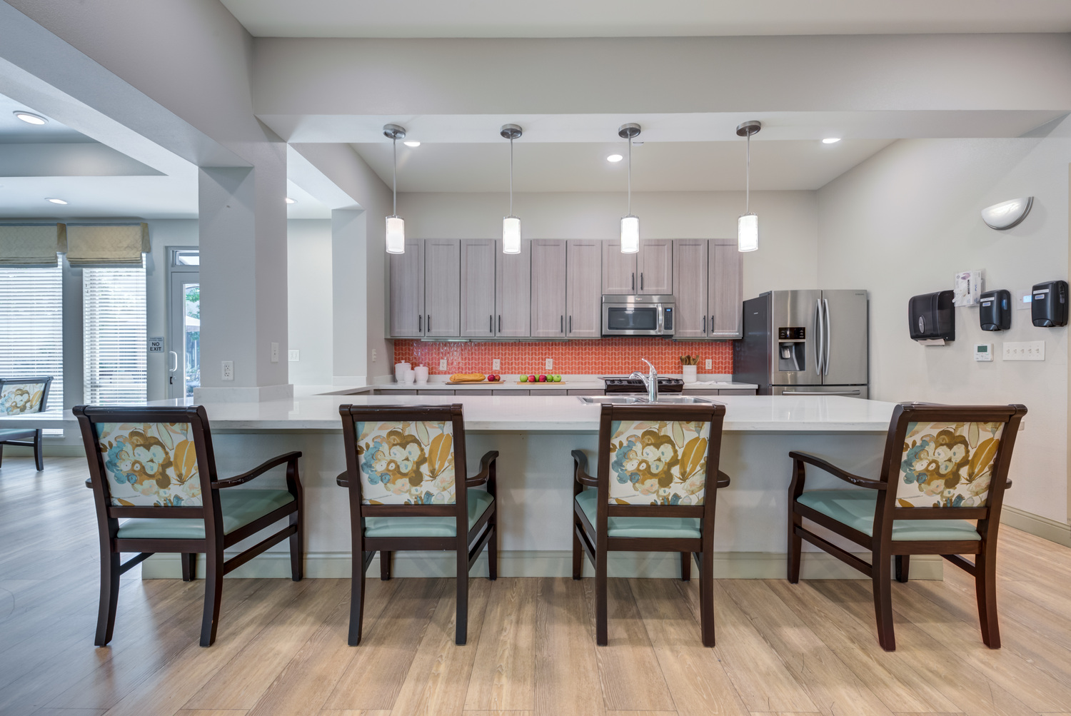 Cozy dining and bar area at this senior living community in Lakeway, TX, with warm wood floors, floral-cushioned chairs, pendant lighting, and an open kitchen in the background.