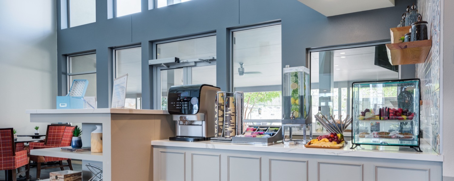 A bright hospitality counter featuring a coffee machine, pod organizer, fresh fruit display, and a glass pastry case, set against large windows and gray cabinetry in a modern lounge or hotel lobby.