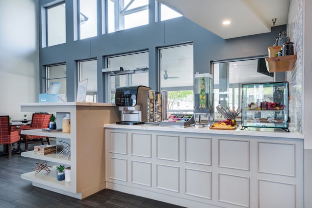 Welcoming bar and café area at this assisted living community in Lakeway, TX, with white cabinetry, open shelving, pendant lighting, and fresh fruits and beverages on display.