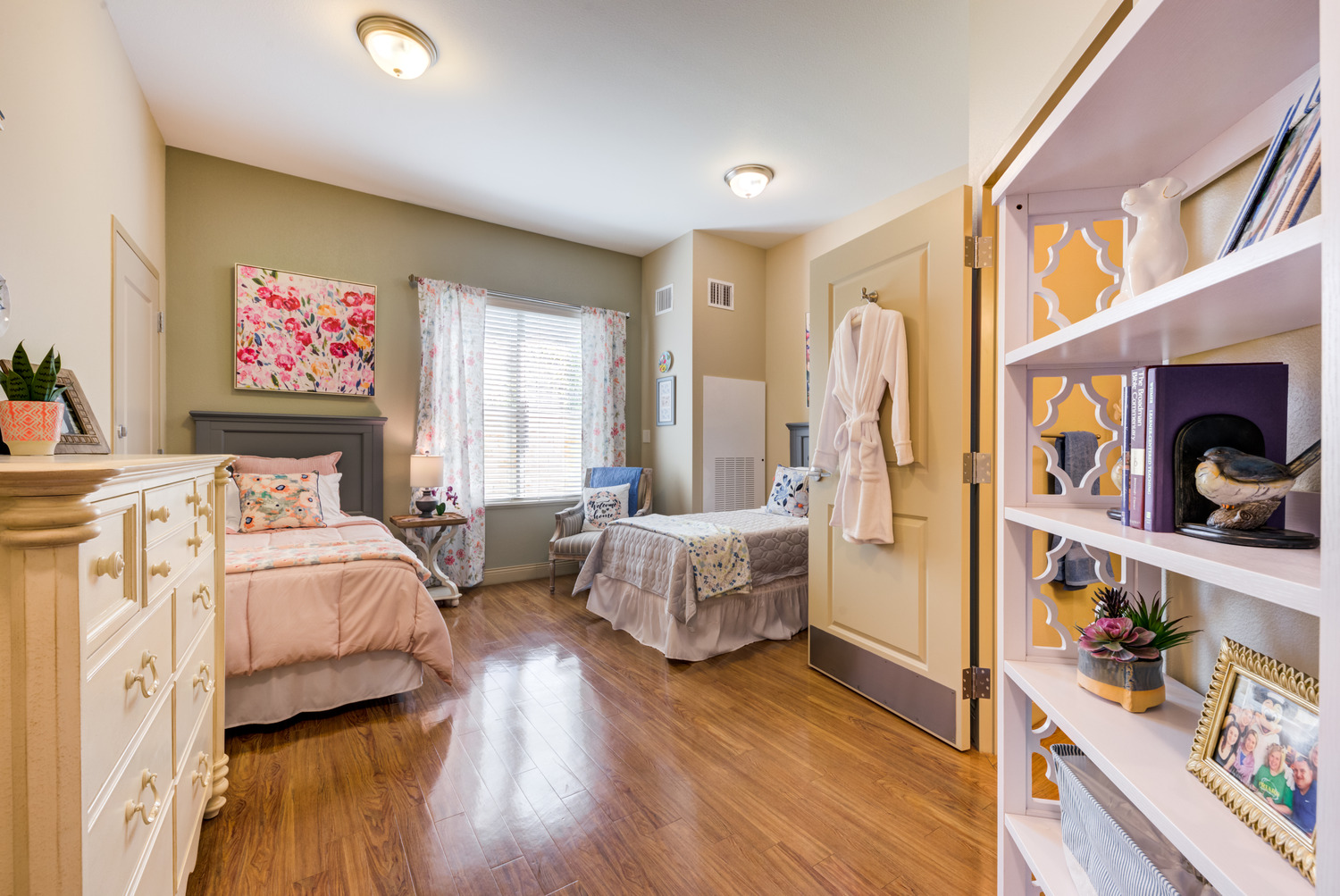 Warmly decorated resident bedroom at Arbor Terrace Lakeway TX assisted living, with wood floors, open shelving, soft bedding, and colorful artwork that gives the space a personalized, homey feel.