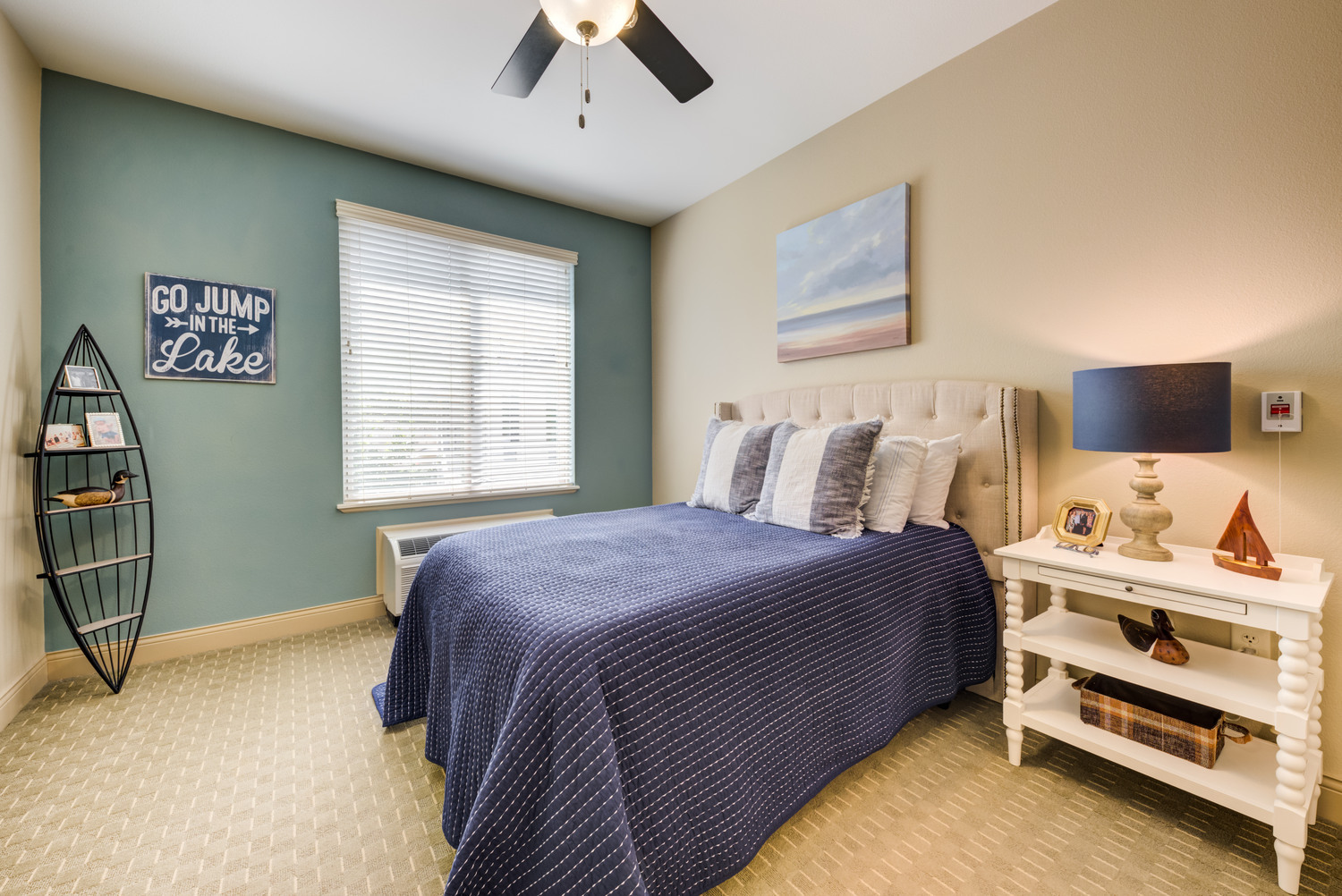 Charming resident bedroom at this senior living community near Lake Travis, featuring a lake-themed accent wall, plush bedding, a ceiling fan, and warm natural light for a relaxing retreat.