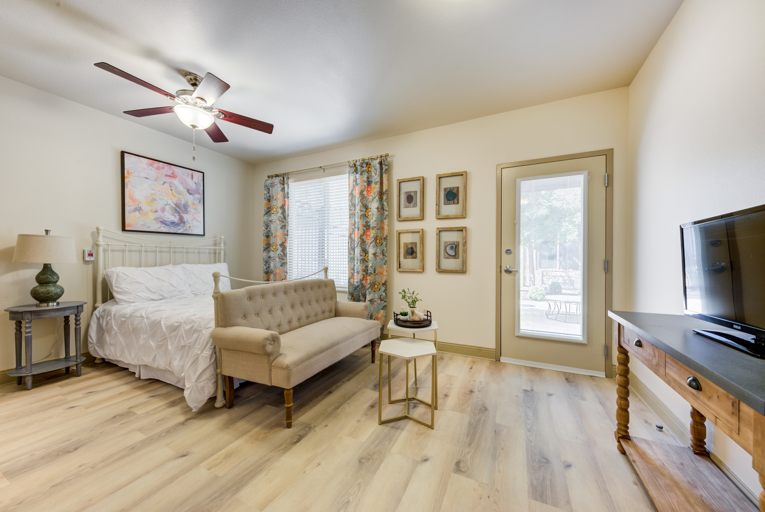 Comfortable and airy resident bedroom at this senior living community in Lakeway, TX, featuring hardwood floors, a ceiling fan, cozy seating area, decorative wall art, and natural light from large windows.