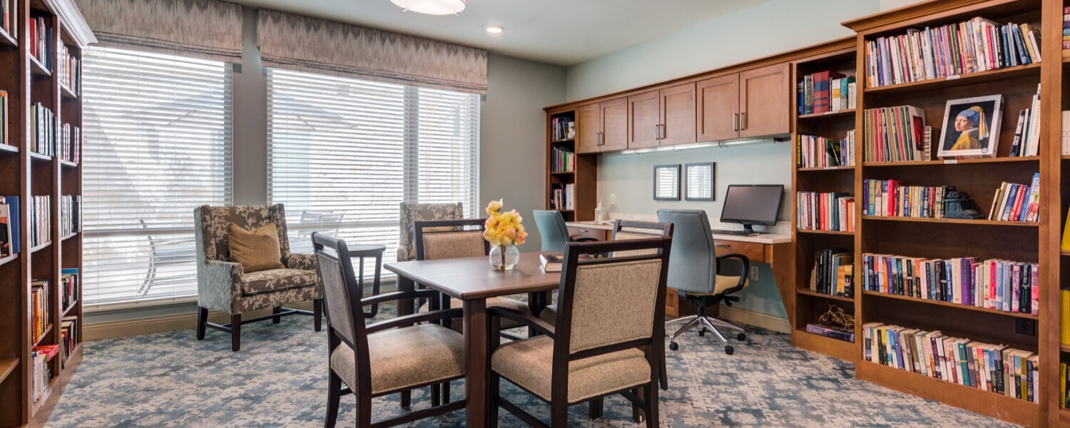 A cozy library and common room featuring a dining table with chairs, two upholstered armchairs near large windows, and built-in wooden bookshelves filled with books along the walls, with a computer workstation in the corner.