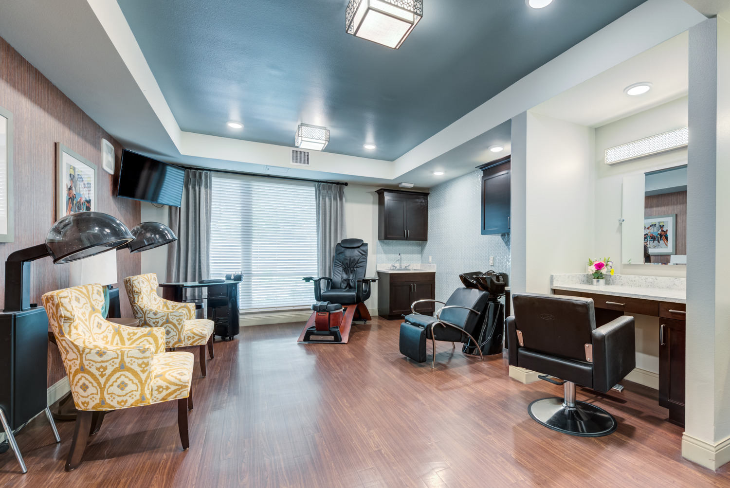 On-site beauty salon featuring salon chairs, a shampoo bowl, hair dryer stations, hardwood flooring, and a tray ceiling with blue accent, offering a full-service salon experience for residents.