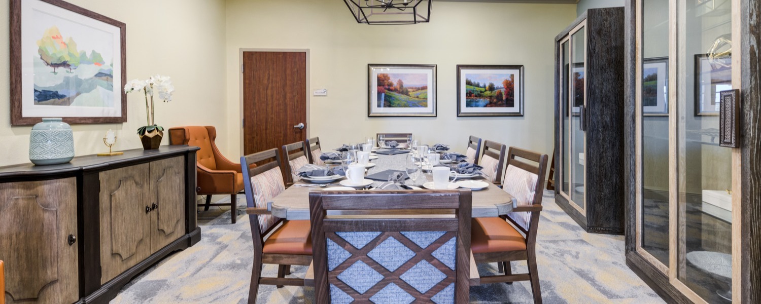 Elegant private dining room with a large round table set for eight, surrounded by wooden chairs with upholstered seats, framed landscape artwork on cream walls, a sideboard with decorative vase and orchid, and a modern geometric light fixture overhead.