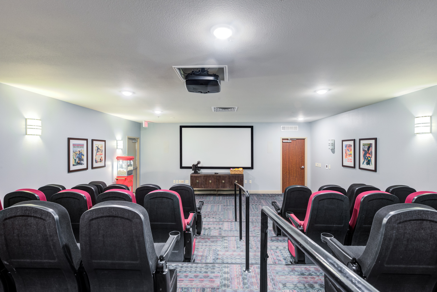 Private resident movie theater with rows of black and red reclining theater seats, a ceiling-mounted projector, a large projection screen, and framed artwork on the walls.