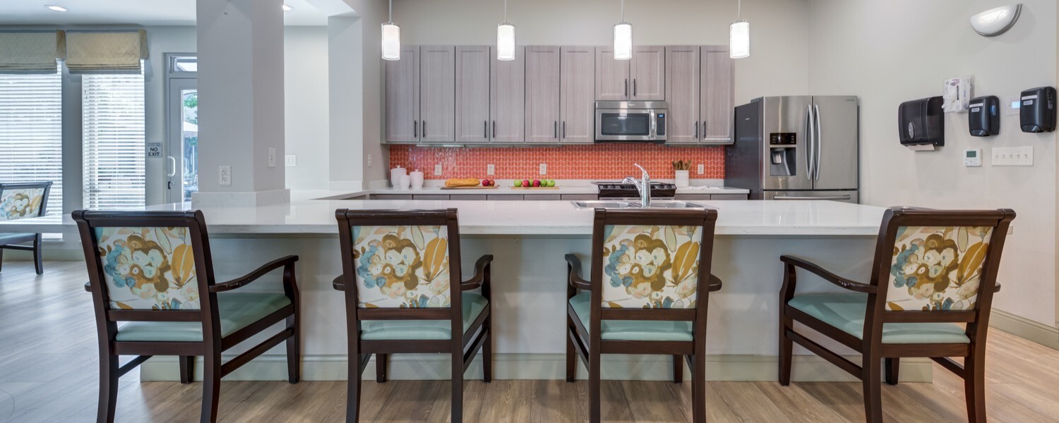 A modern kitchen and dining area featuring a large white quartz island with four upholstered chairs in a floral and teal pattern, gray cabinetry, a red tile backsplash, stainless steel appliances, and pendant lighting over light hardwood flooring.