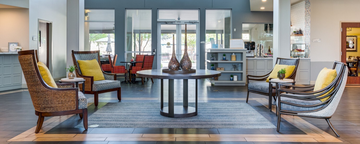 Elegant and inviting lobby at Arbor Terrace Lakeway TX senior living, with contemporary furnishings, yellow accent chairs, a round wood table, and an open layout filled with natural light.