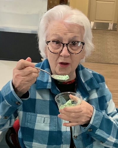 An older woman with white hair and glasses enjoys a spoonful of green ice cream or sorbet from a clear plastic cup, wearing a blue plaid shirt.