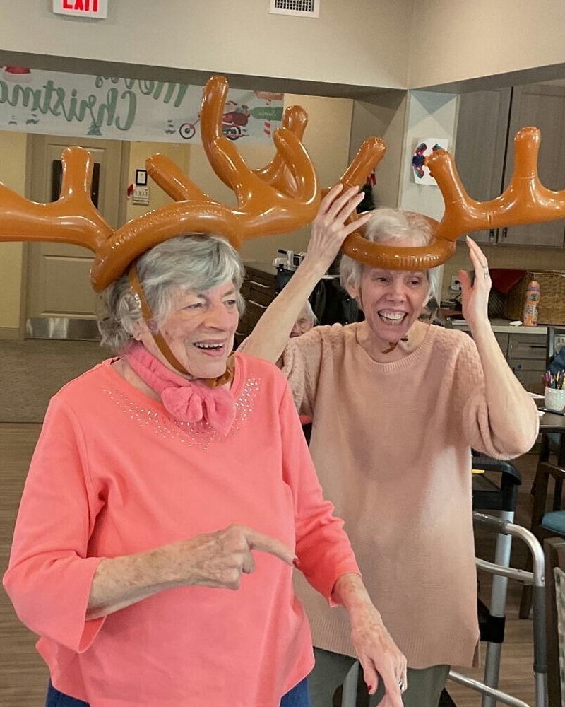 Two elderly women wearing inflatable reindeer antler headbands laugh and smile together during a festive holiday activity.