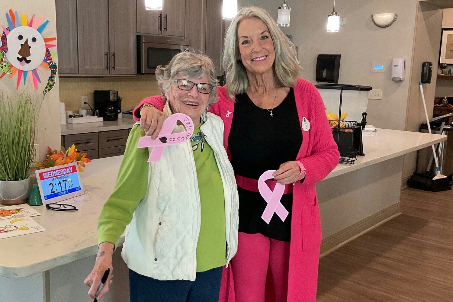 A resident and a staff member smile together while holding pink ribbons in support of Breast Cancer Awareness at Arbor Terrace Lakeway, a trusted assisted living community in Lakeway, TX.