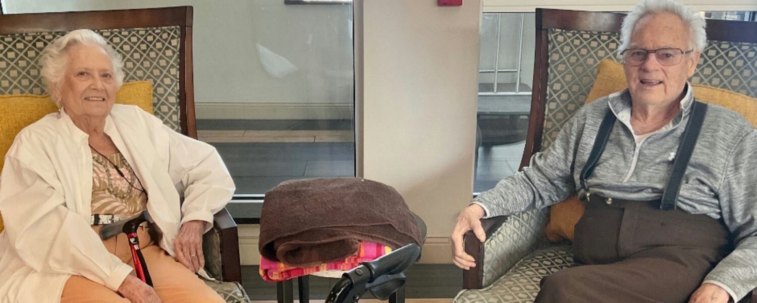 Two older adults sit smiling in comfortable armchairs by a bright window, with a bag and hat resting on a table between them, enjoying a relaxed visit together.