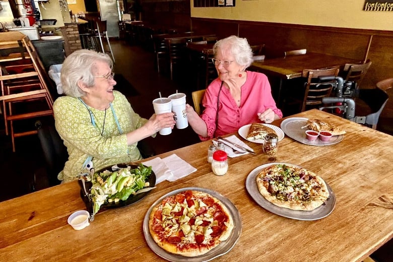 Two residents share a cheerful toast over a shared pizza meal at this senior living community in Lakeway, TX, enjoying a fun and relaxed dining experience together at Arbor Terrace.