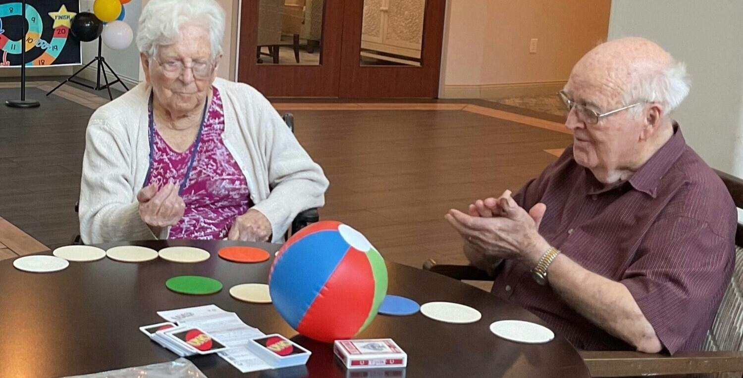 Two elderly residents sit across from each other at a table, engaged in a colorful disc and beach ball activity, with playing cards and colored circles spread across the table between them.