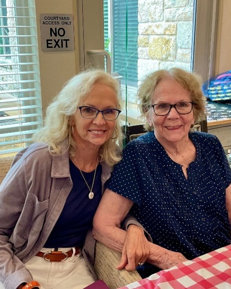 Two women sitting closely together and smiling at the camera, one with long blonde hair wearing a gray cardigan, and the other with short white hair wearing a navy blue polka-dot top.