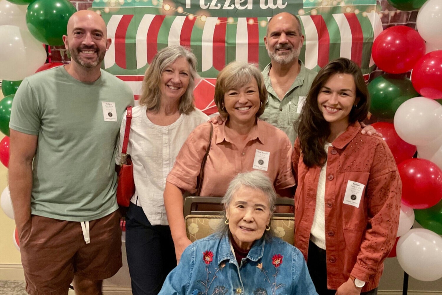 A group of five adults and one elderly woman pose together smiling in front of a festive pizzeria-themed backdrop decorated with red, white, and green balloons.