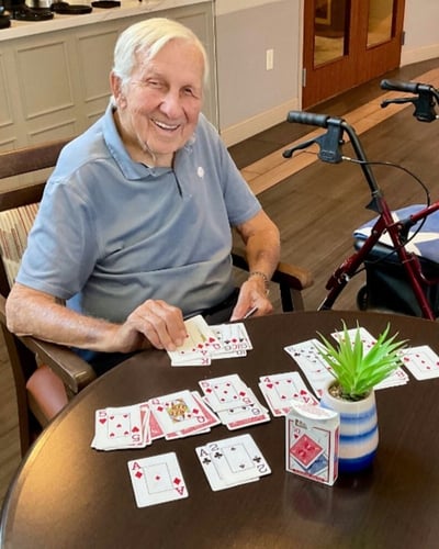 A smiling older man sits at a wooden table playing a card game, with multiple playing cards spread out in front of him and a small potted plant nearby, in a warmly lit room.