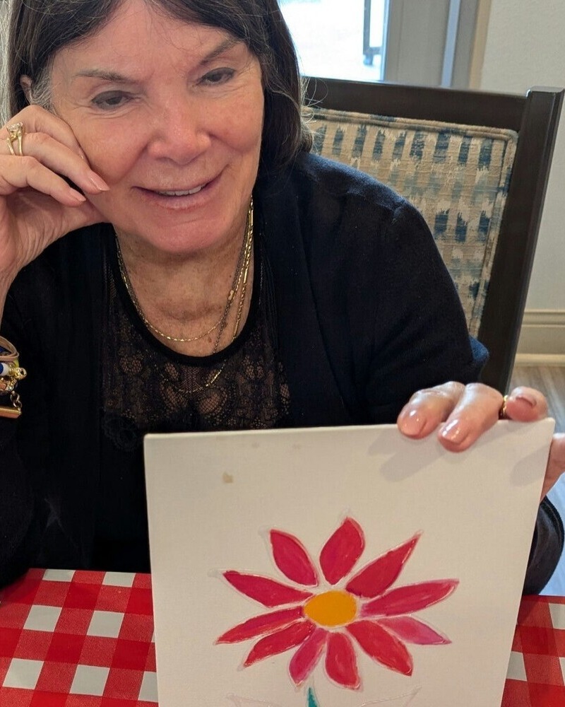 A smiling woman holds up a painting she created of a red flower with a yellow center, sitting at a table with a red checkered tablecloth during an art activity.