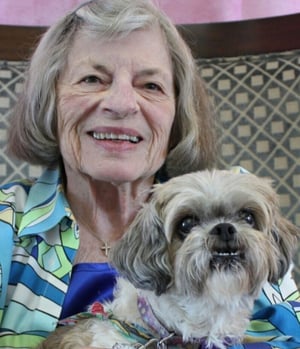 An elderly woman with short gray hair smiles warmly while holding a small Shih Tzu dog. She is wearing a colorful floral blouse and a blue necklace.