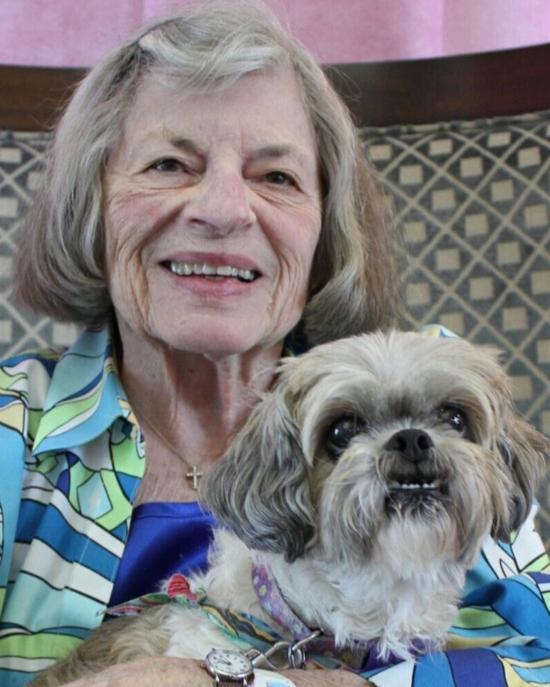 An elderly woman with short gray hair smiles warmly while holding a small Shih Tzu dog. She is wearing a colorful floral blouse and a blue necklace.