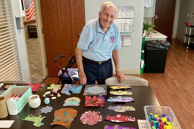 An older man stands proudly behind a table displaying colorful handmade craft projects in a senior living community activity room.
