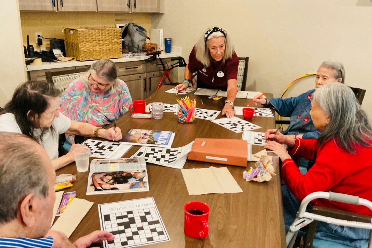 A group of seniors and a staff member gather around a table working on large-print crossword puzzles together in a cozy activity room.