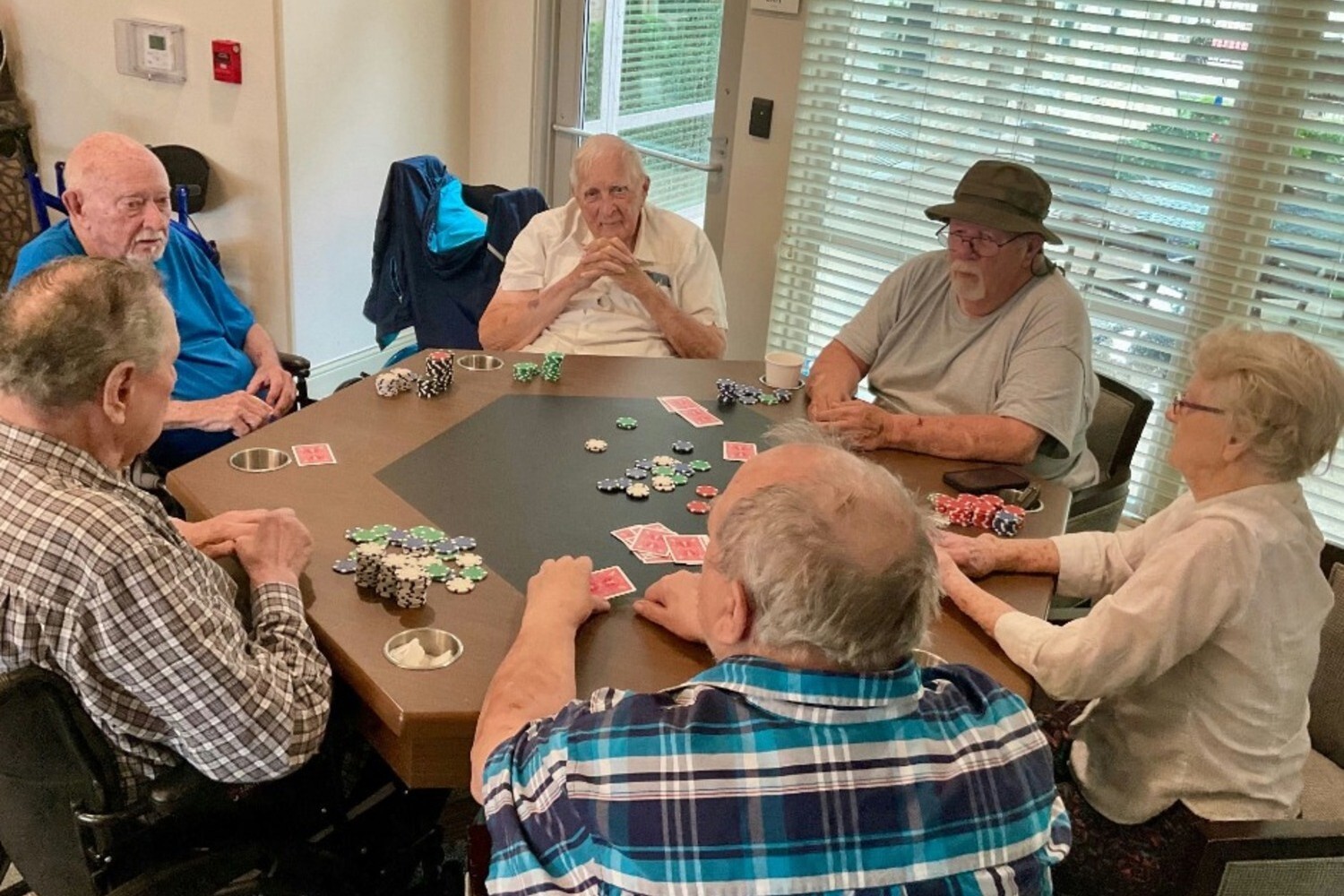 A group of six older adults gathered around a round table playing poker, with playing cards and poker chips spread across the table, in a bright common room with large windows.